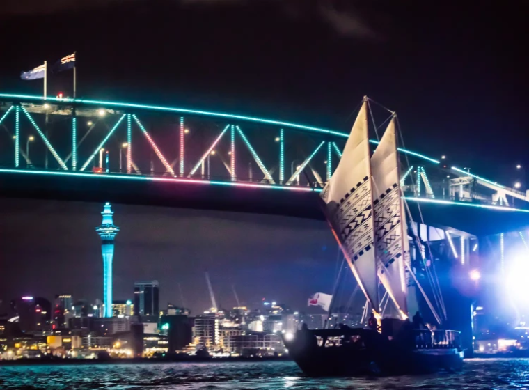 Auckland Harbour Bridge lit up at night with a double-hulled canoe on the water.