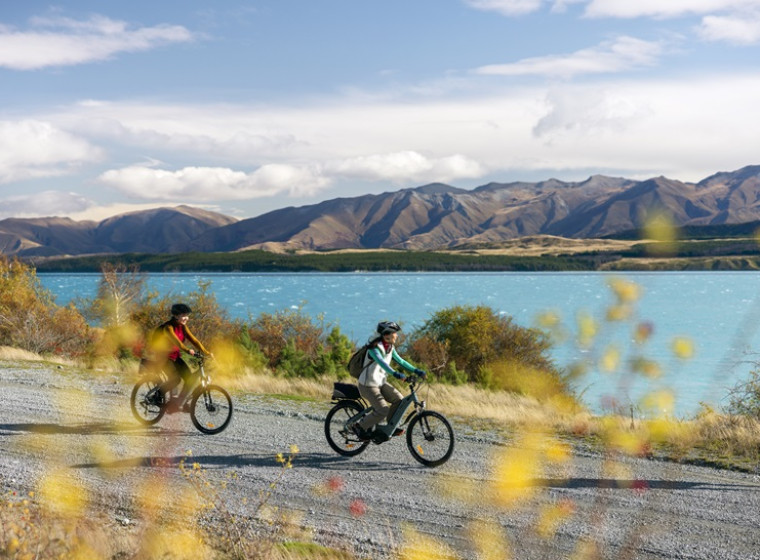 2 people cycle along a gravel track beside Lake Tekapo in the Autumn.